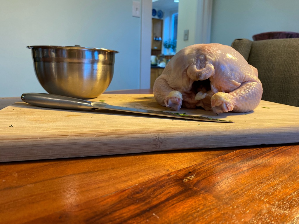Image of a raw chicken, a knife, and a bowl sitting on a wooden cutting board. 
