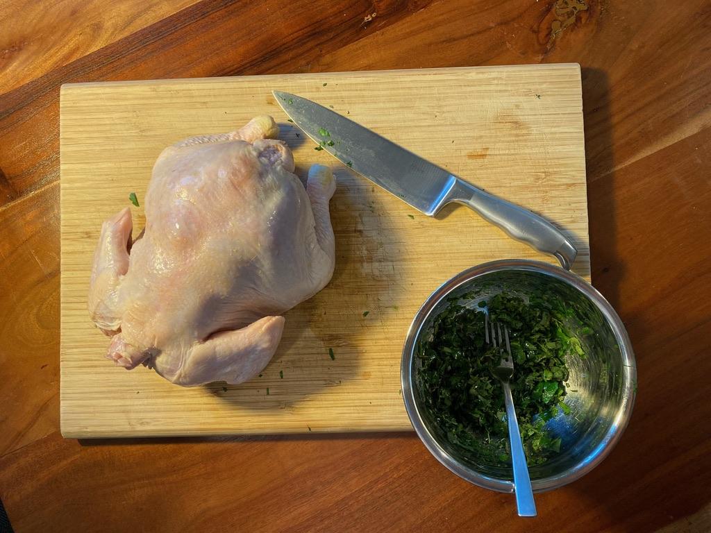 Image of a raw chicken, knife, and bowl sitting on a cutting board. Inside the bowl are herbs and a fork.